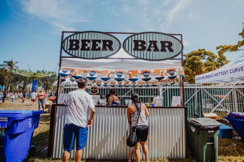A lively beer bar at an outdoor event, with people ordering drinks under a sunny sky.