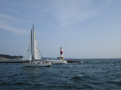 Sailboats on a calm sea near a pier with a red and white lighthouse under a clear blue sky.