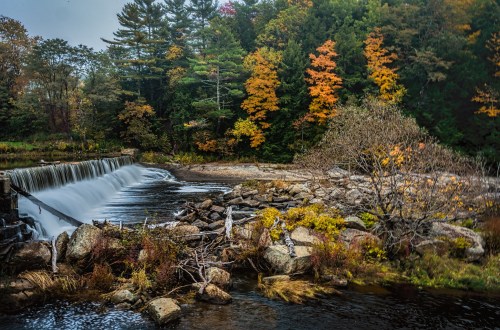 A serene waterfall cascades over rocks, surrounded by vibrant autumn foliage and a tranquil riverbank.