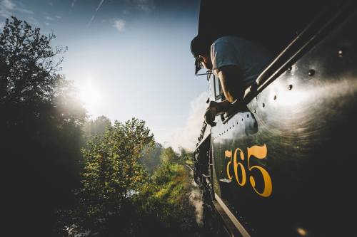 A vintage steam train chugs along a scenic route, with a passenger leaning out the window, surrounded by greenery.