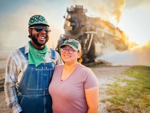 A smiling couple stands in front of a steam train, with smoke billowing in the background during sunset.