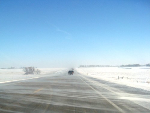A snowy road stretches into the distance under a clear blue sky, with strong winds creating a blizzard effect.