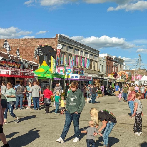 A busy street fair with colorful tents, food stalls, and crowds of people enjoying the festivities under a blue sky.