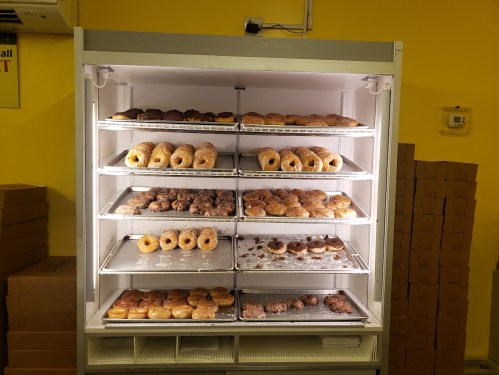 A display case filled with various types of donuts and pastries on multiple shelves, illuminated against a yellow background.