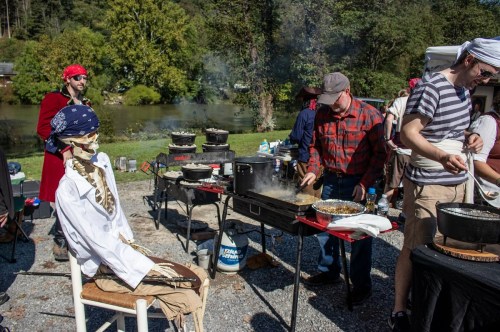 A lively outdoor cooking scene with people preparing food, a skeleton in a chef's outfit sitting nearby.