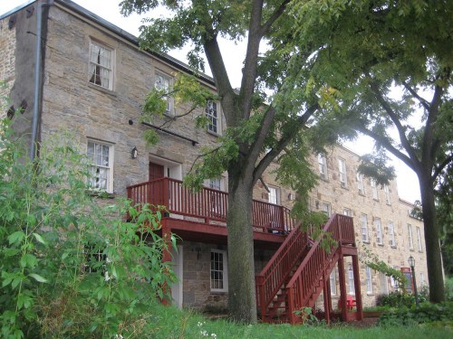 Historic stone building with a wooden staircase and balcony, surrounded by trees and greenery.