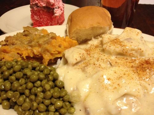 A plate of comfort food featuring biscuits and gravy, green peas, sweet potato casserole, a roll, and dessert.