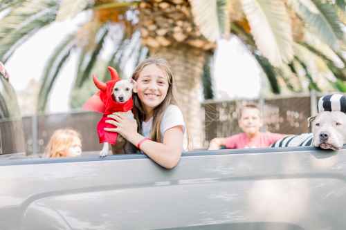 A girl in a red costume holds a small dog, smiling, with two other kids and a dog in the background near palm trees.