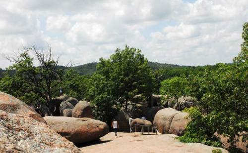 A scenic landscape featuring large boulders, lush greenery, and a person walking under a partly cloudy sky.
