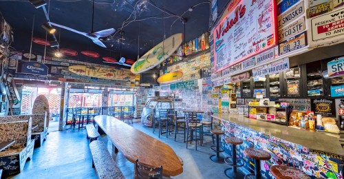 A vibrant bar interior with surfboards, colorful stickers, and a long communal table, creating a lively atmosphere.