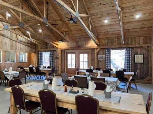 A rustic dining hall with wooden beams, tables set for meals, and large windows letting in natural light.