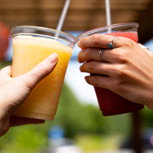 Two hands clink cups of yellow and red frozen drinks, with a sunny outdoor background.