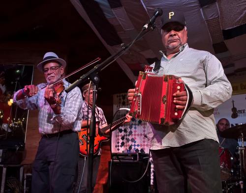Musicians perform on stage, one playing the violin and another the accordion, with a guitar player in the background.