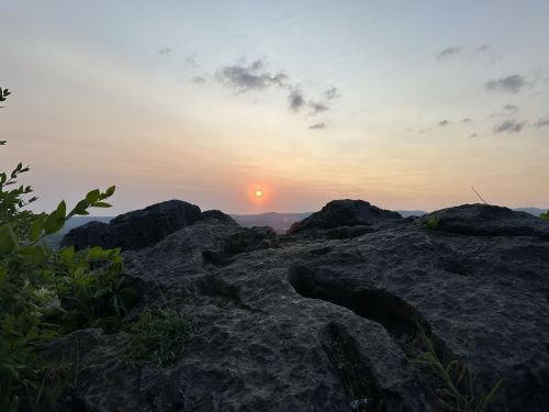 A sunset view from a rocky ledge, with soft clouds and distant mountains in the background.