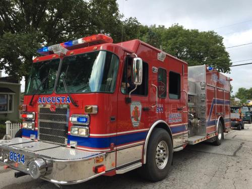 A red fire truck with lights flashing, parked on a street surrounded by trees and buildings.