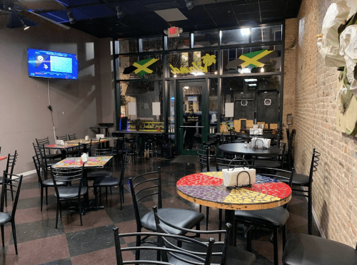Interior of a restaurant with colorful tables, black chairs, and a TV, featuring Jamaican decor and large windows.