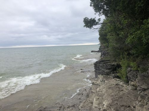 A rocky shoreline meets a calm lake under a cloudy sky, with trees lining the edge. Waves gently lap at the rocks.