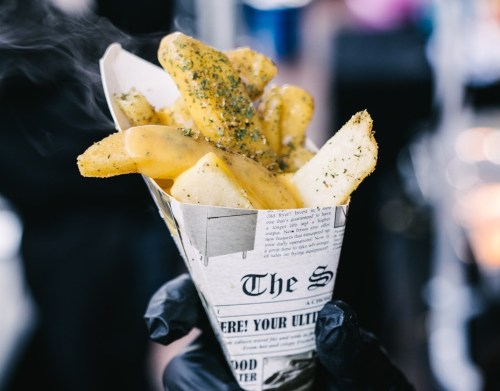 A hand holding a cone of golden fries topped with melted cheese and herbs, served in a newspaper-style wrapper.