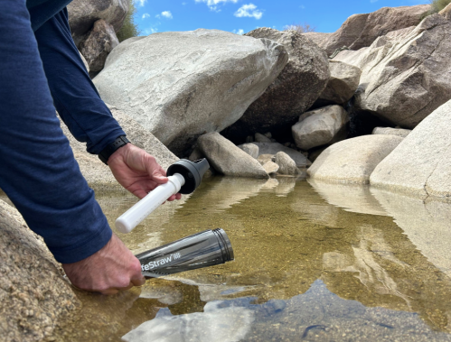 A person uses a water filter straw to collect water from a shallow pool surrounded by rocks.