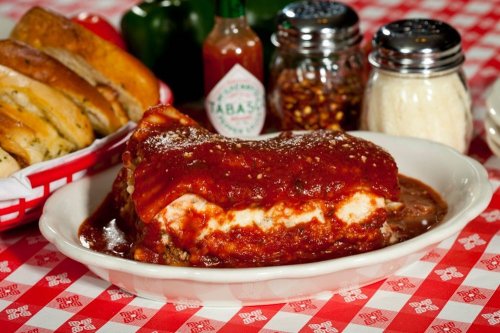 A plate of lasagna topped with marinara sauce, surrounded by condiments and garlic bread on a checkered tablecloth.