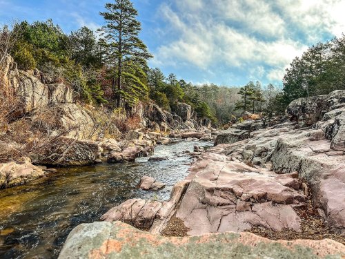 A serene river flows through rocky terrain, surrounded by trees and a cloudy blue sky.