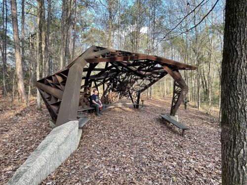 A unique, modern shelter made of metal beams in a wooded area, with two people sitting on benches inside.