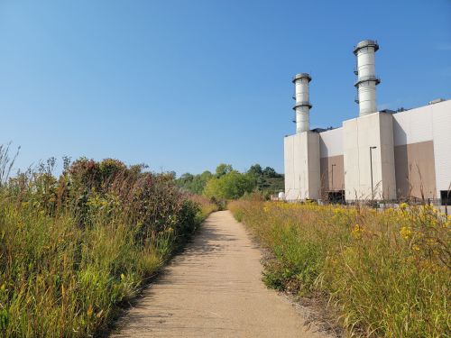 A sandy path leads through tall grass toward industrial buildings with smokestacks under a clear blue sky.