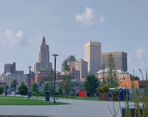 City skyline featuring modern and historic buildings under a clear blue sky, with green park space in the foreground.