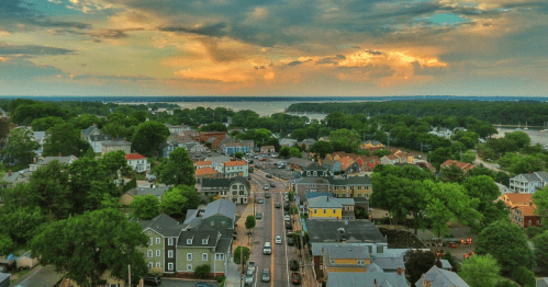 Aerial view of a charming town with colorful houses, trees, and a river under a dramatic sunset sky.