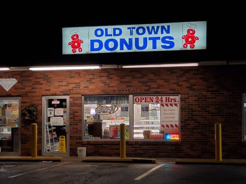 A brightly lit donut shop at night with a sign reading "Old Town Donuts" and a window displaying various treats.