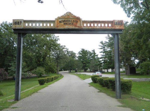 Archway sign for Nebraska City Bridge, flanked by trees and a pathway, with a car in the distance.