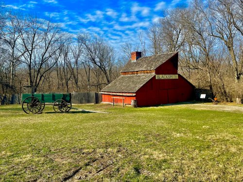 A red barn with a sign reading "Blacksmith" beside a green wagon, set in a grassy area with bare trees and blue sky.