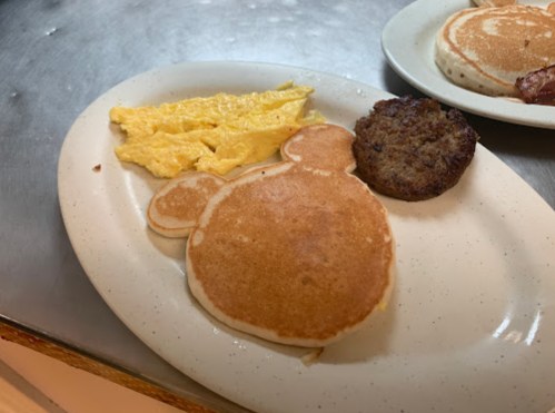 A plate with a bear-shaped pancake, scrambled eggs, and a sausage patty.