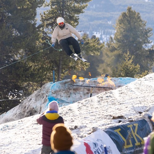 A skier jumps off a snowy ramp, with flames erupting below, while spectators watch from the side.