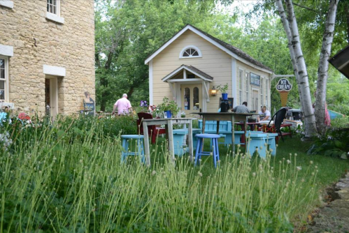 A quaint outdoor scene featuring colorful chairs and tables surrounded by greenery, with a charming house in the background.