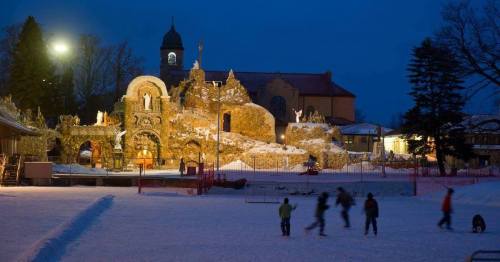A snowy landscape at dusk featuring a large, illuminated ice sculpture and people skating nearby.