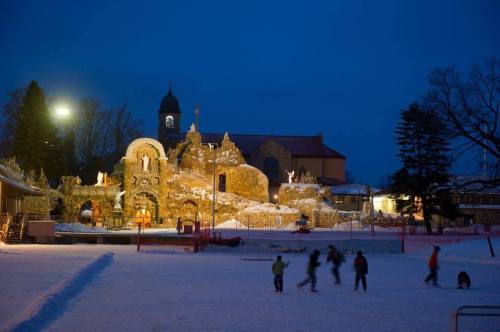 A snowy scene at dusk with a historic building and people skating on an ice rink in the foreground.