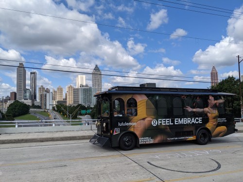A Lululemon-branded trolley bus on a city street with a skyline of tall buildings and a blue sky in the background.