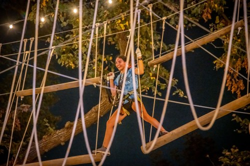 A person navigates a high ropes course at night, smiling as they walk on wooden beams surrounded by trees and lights.
