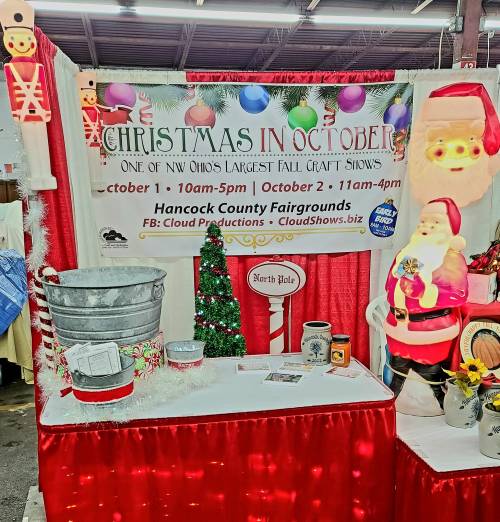 A festive booth at a craft show featuring a Santa figure, holiday decorations, and a sign for "Christmas in October."