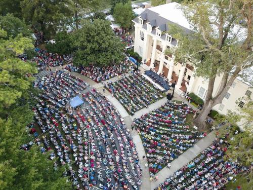Aerial view of a large crowd seated in an outdoor amphitheater near a historic building, surrounded by trees.