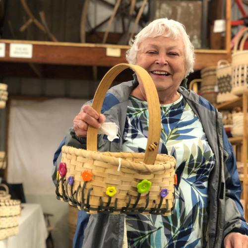 Smiling older woman holding a colorful woven basket with flowers, standing in a market filled with baskets.