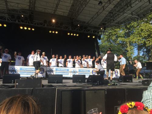 A group of performers in matching shirts on stage at an event, with a crowd and trees in the background.