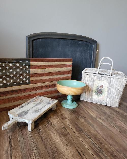 A rustic display featuring a wooden American flag, a bowl on a pedestal, a woven basket, and a cutting board on a wooden table.
