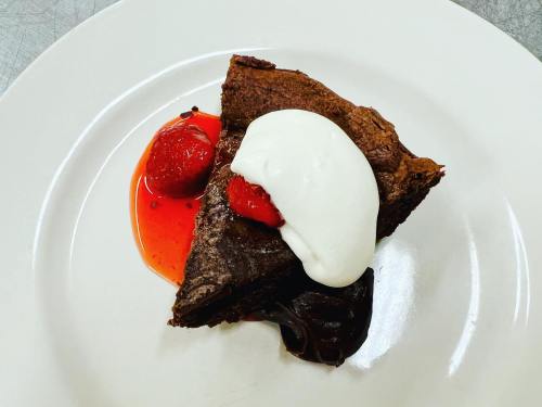 A slice of chocolate cake topped with whipped cream and strawberries, served on a white plate with berry sauce.