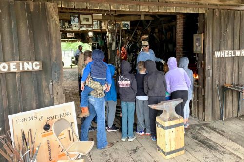 A group of people gathers around a blacksmith demonstrating tools in a rustic workshop.