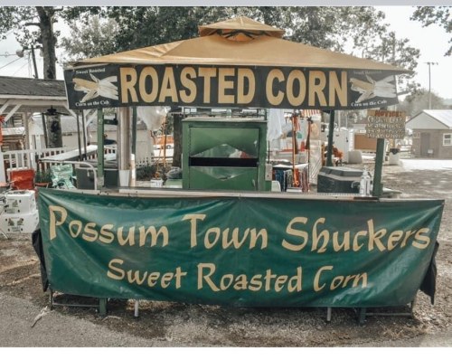 A food stand with a yellow roof, featuring a sign for "Roasted Corn" and a green banner for "Possum Town Shuckers."