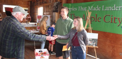 A man hands a card to a young couple at an apple festival in a rustic barn setting. Green banners decorate the background.