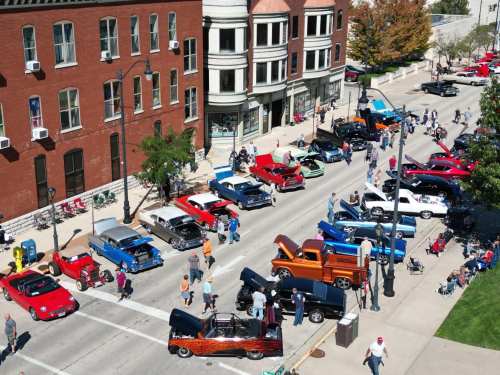 Aerial view of a car show featuring classic cars, with people gathered along the street and buildings in the background.