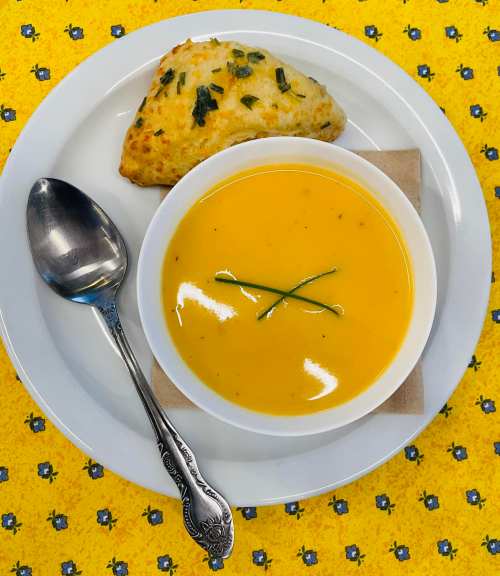 A bowl of creamy yellow soup garnished with herbs, accompanied by a scone on a white plate and a silver spoon.